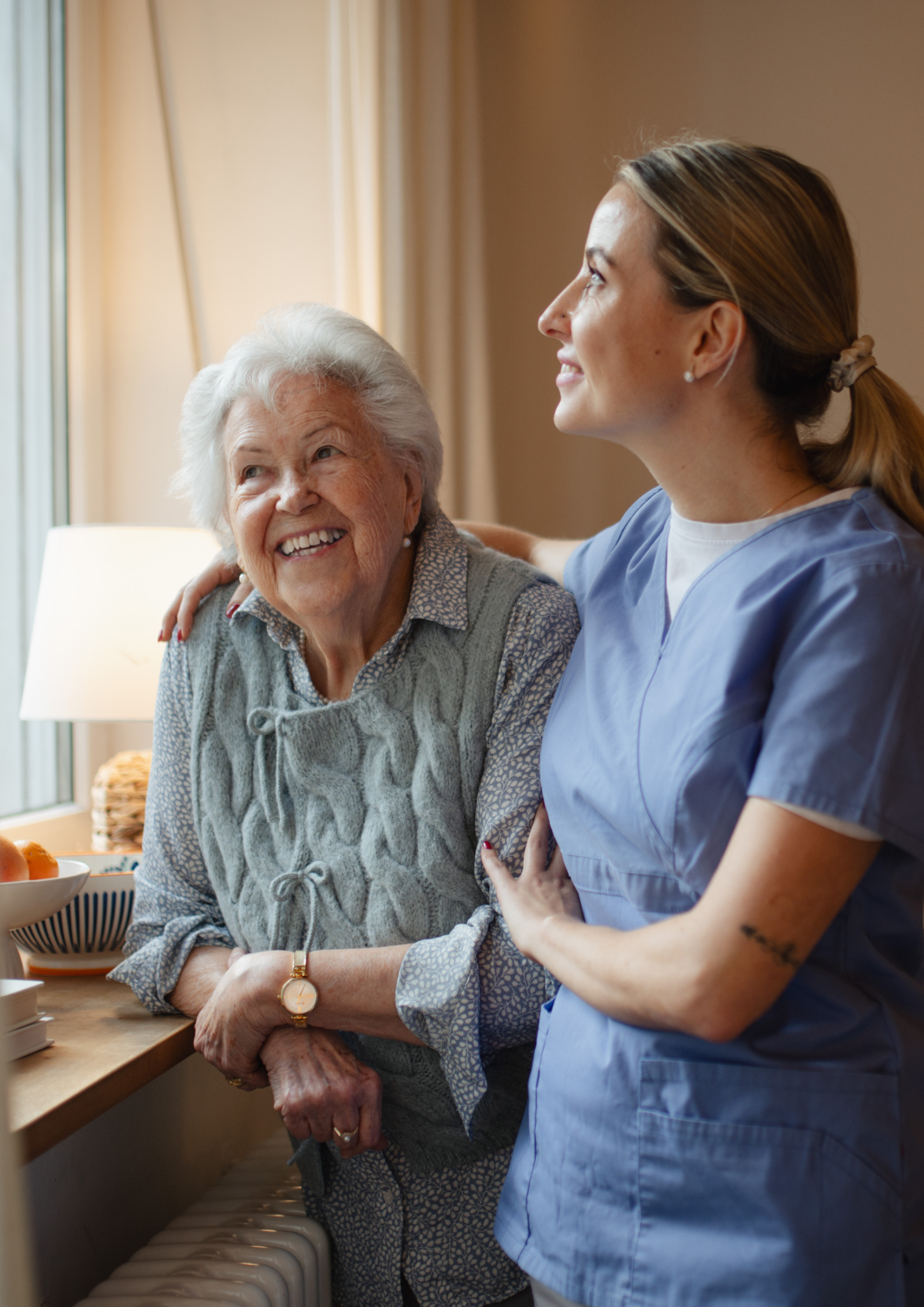 Smiling elderly woman with arm around her is comforted by a caregiver by a window.