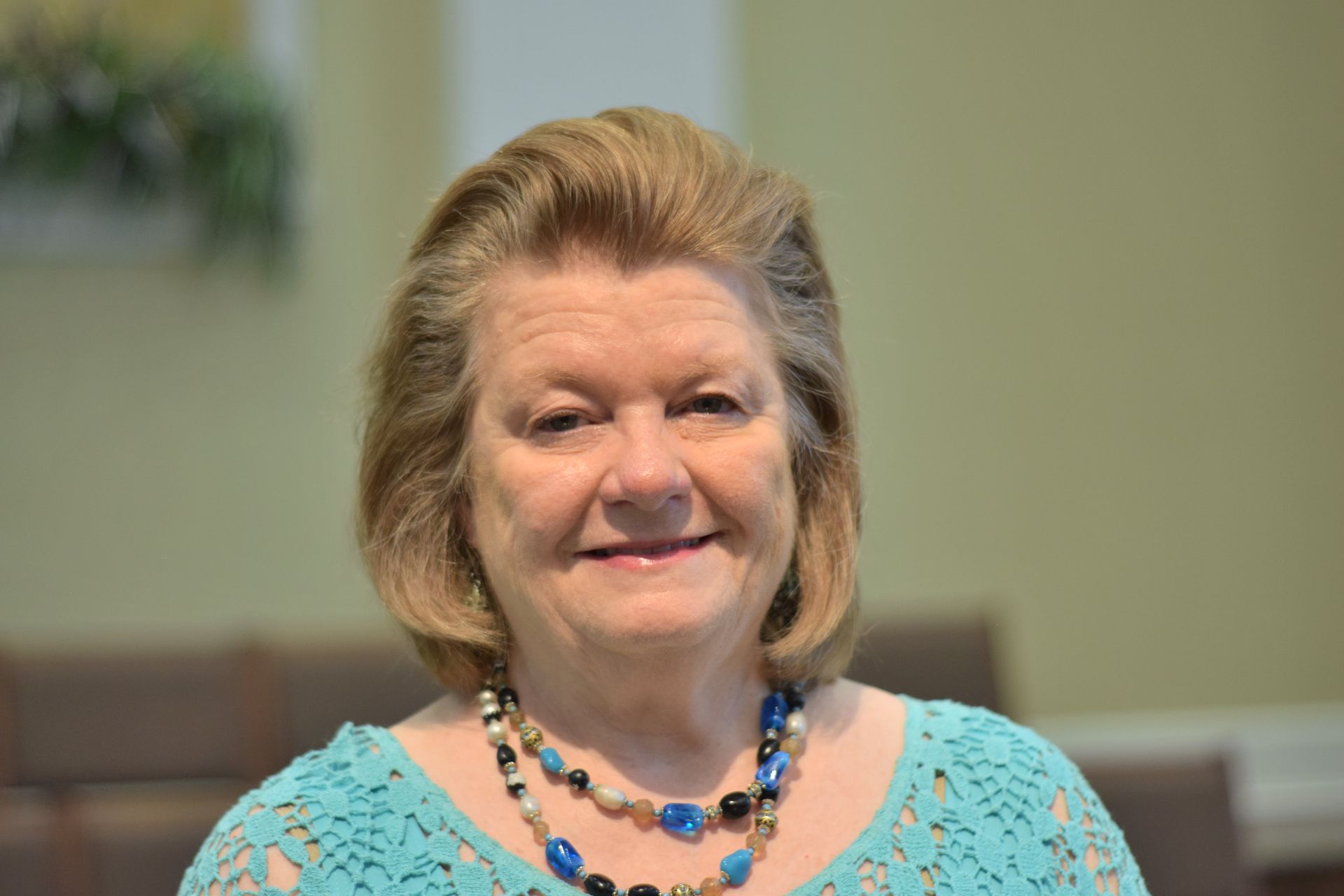 Woman smiling in a turquoise top and beaded necklace, seated indoors against a blurred background