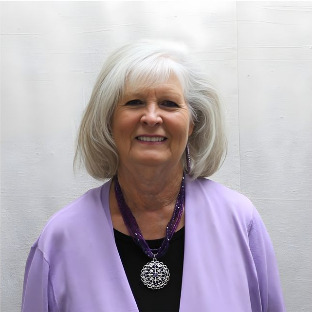 Smiling woman with white hair wearing a lavender cardigan and black top against a light background