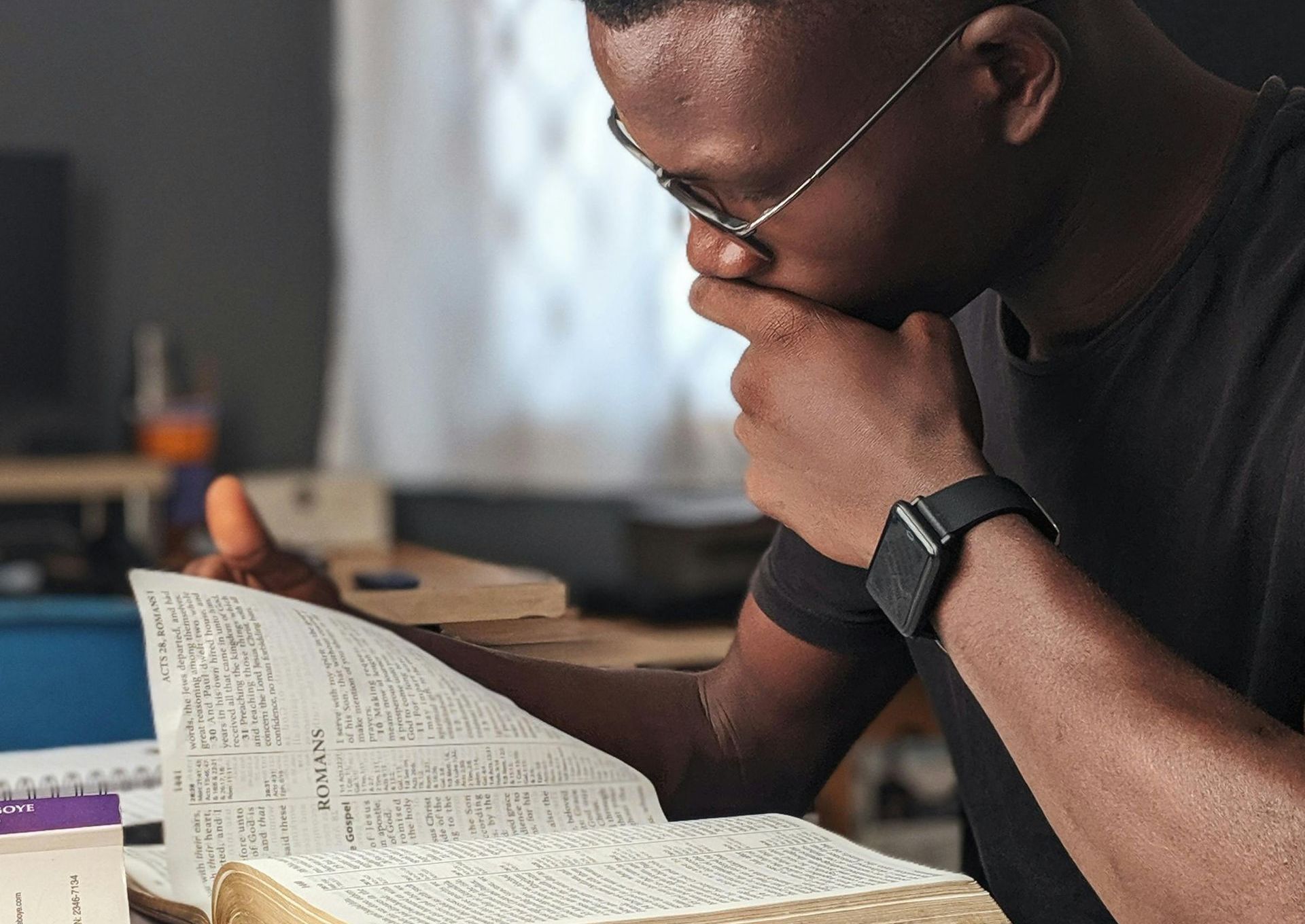 Person in glasses studies a book, hand to chin, indoors.