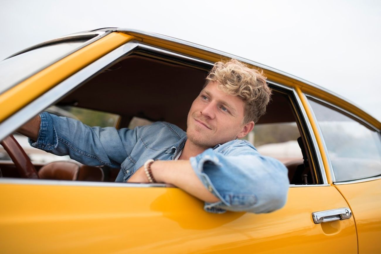 Man with curly hair looking out of the window of a vintage yellow car.