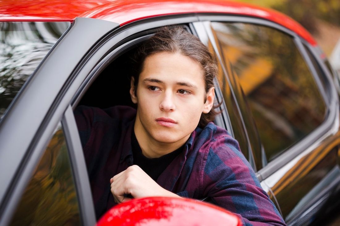 Teen with a serious expression looking out from the driver's side window of a red car.