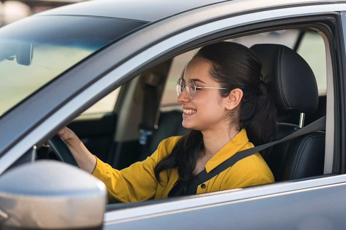 A woman wearing glasses and a yellow shirt smiling while driving a car.