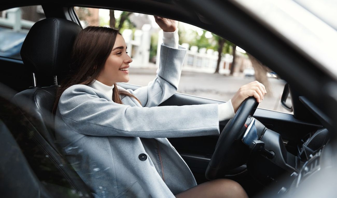 A smiling woman driving a car and adjusting the rearview mirror.