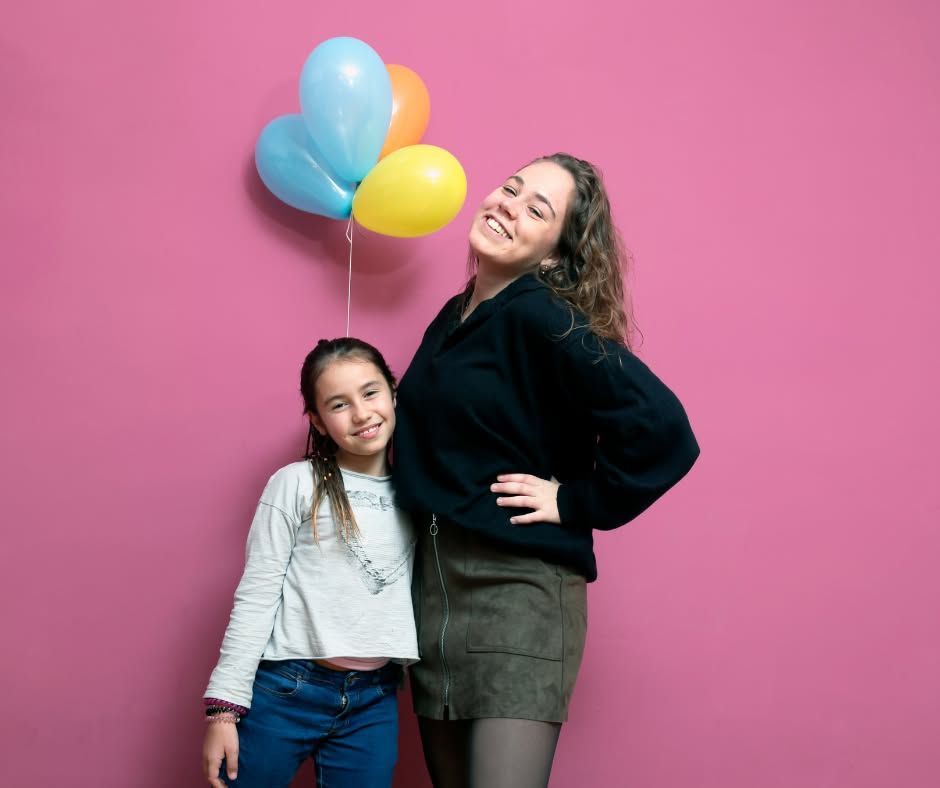 Two people posing with colorful balloons against a pink wall, smiling at the camera.
