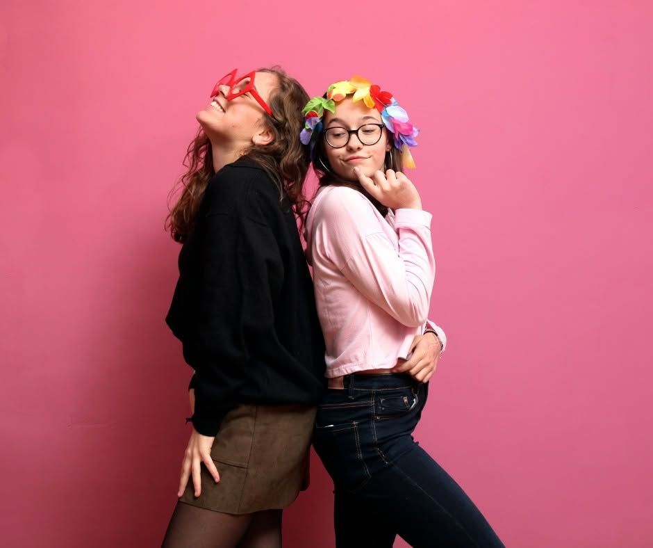 Two friends posing back-to-back against a pink background, wearing colorful flower crowns and smiling.