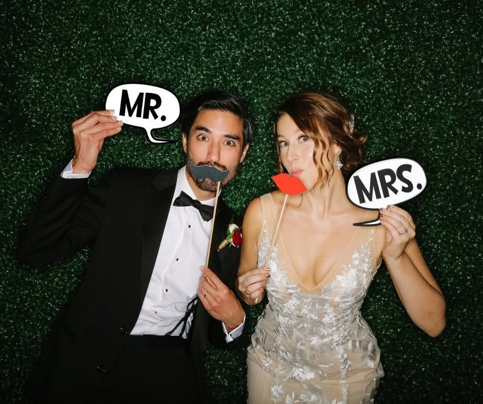 Two people in formal wear holding “MR.” and “MRS.” signs at a sparkly party backdrop