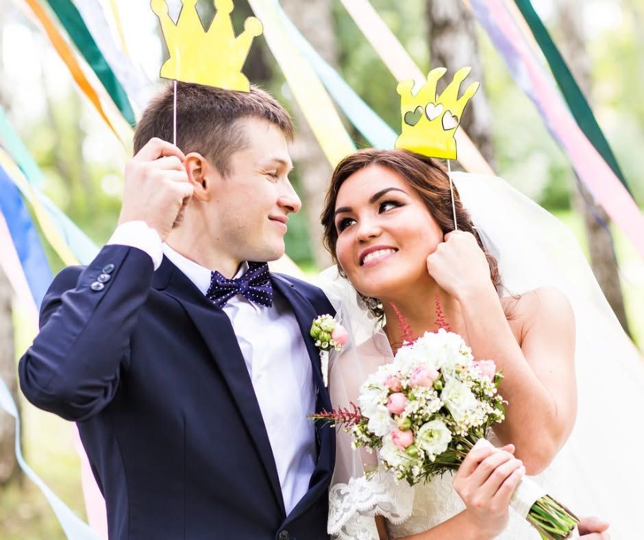 Bride and groom smiling with paper crowns and bouquet at a wedding celebration