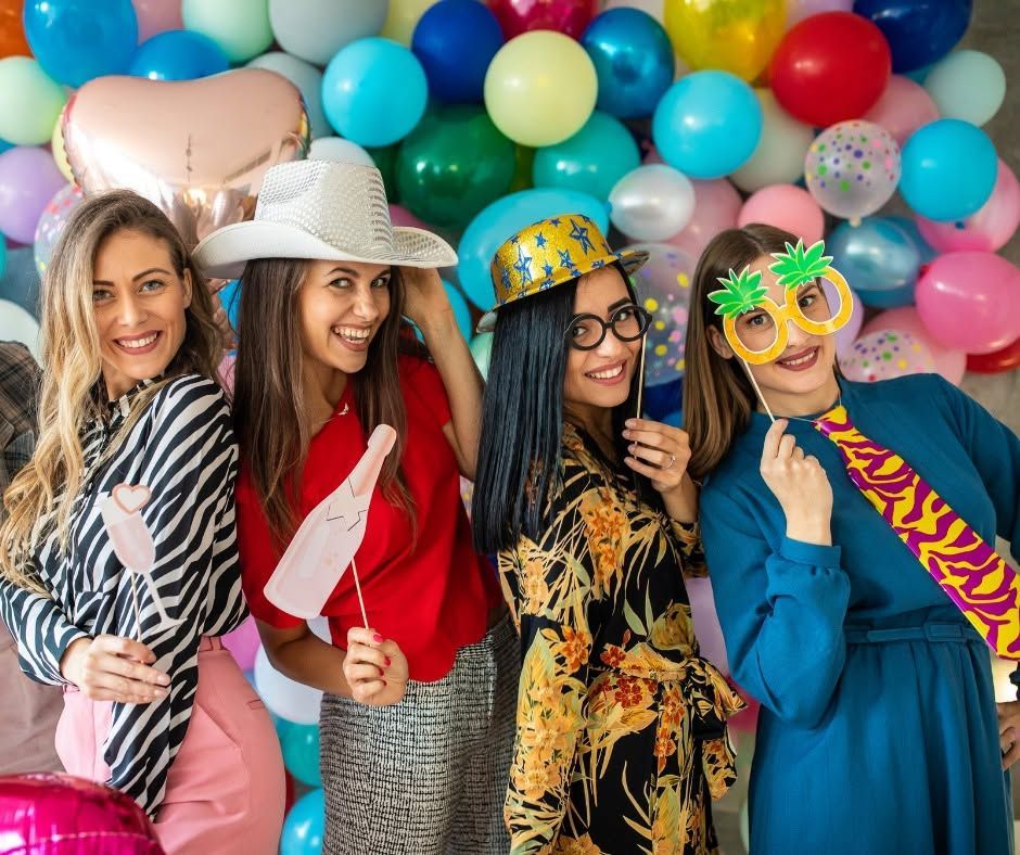 Four friends posing at a colorful party, holding props and wearing festive accessories.