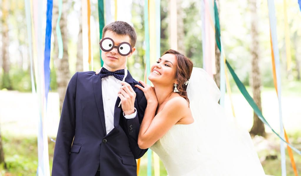 Bride laughing beside groom in suit at an outdoor wedding, with colorful ribbons in the background