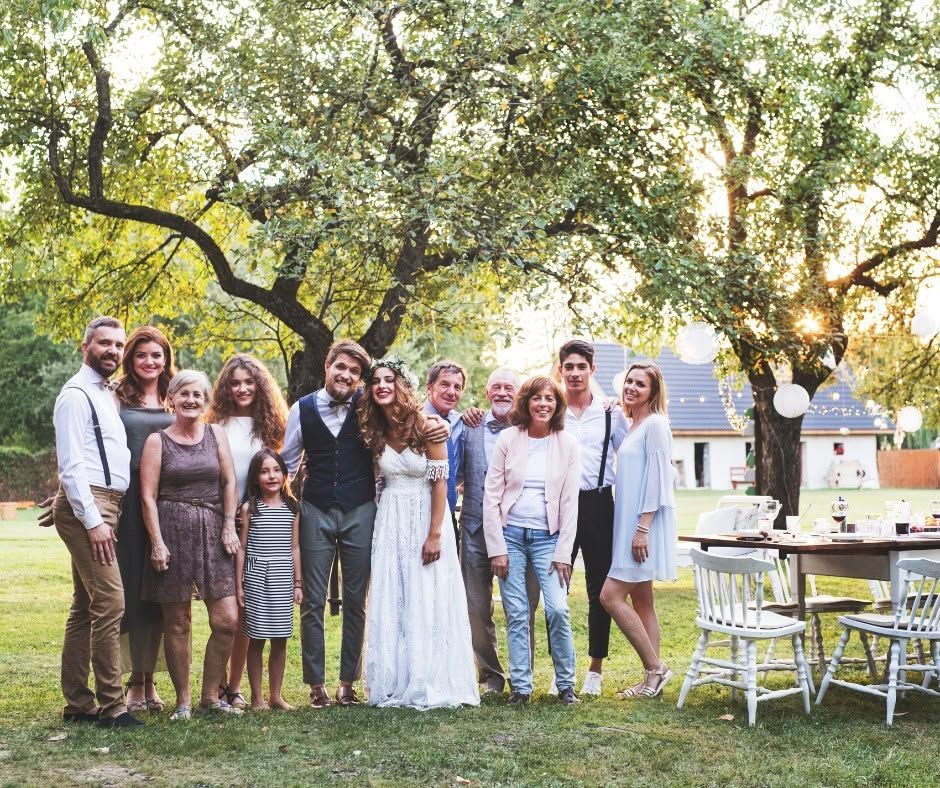 Large family group standing together outdoors in a sunny backyard with a decorated table and white chairs