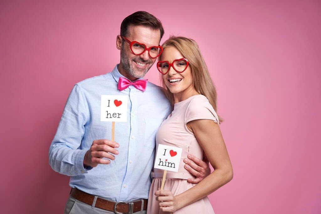 Two people in red glasses and bow ties hold mugs against a pink studio backdrop.