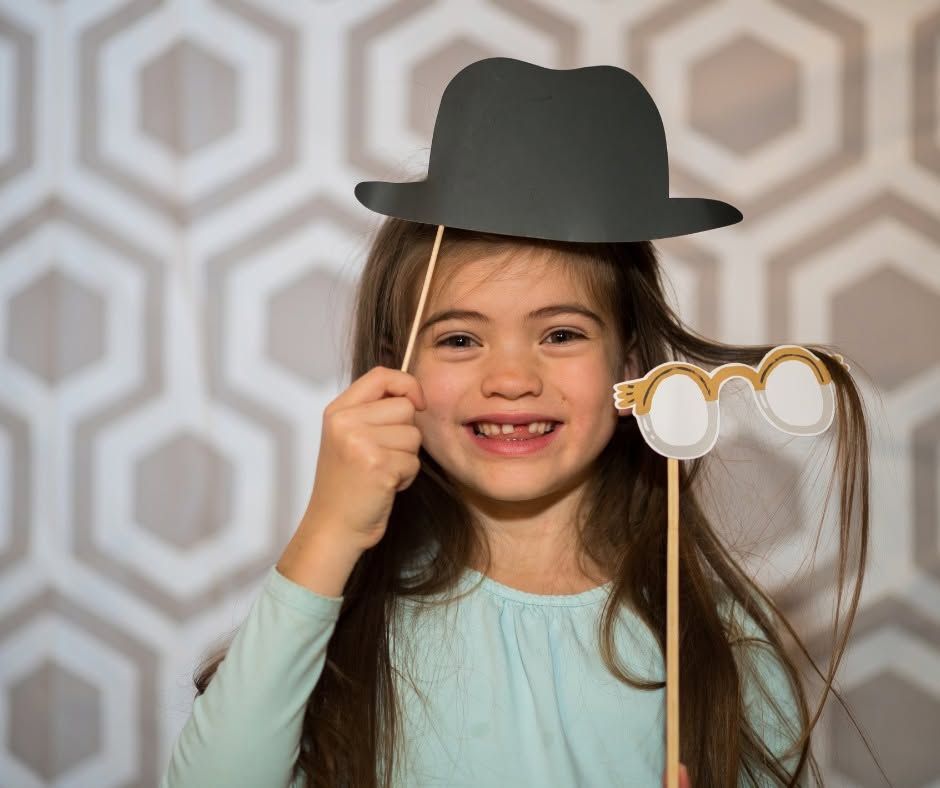 Smiling child holding a gray hat and round glasses, standing against a patterned wall.