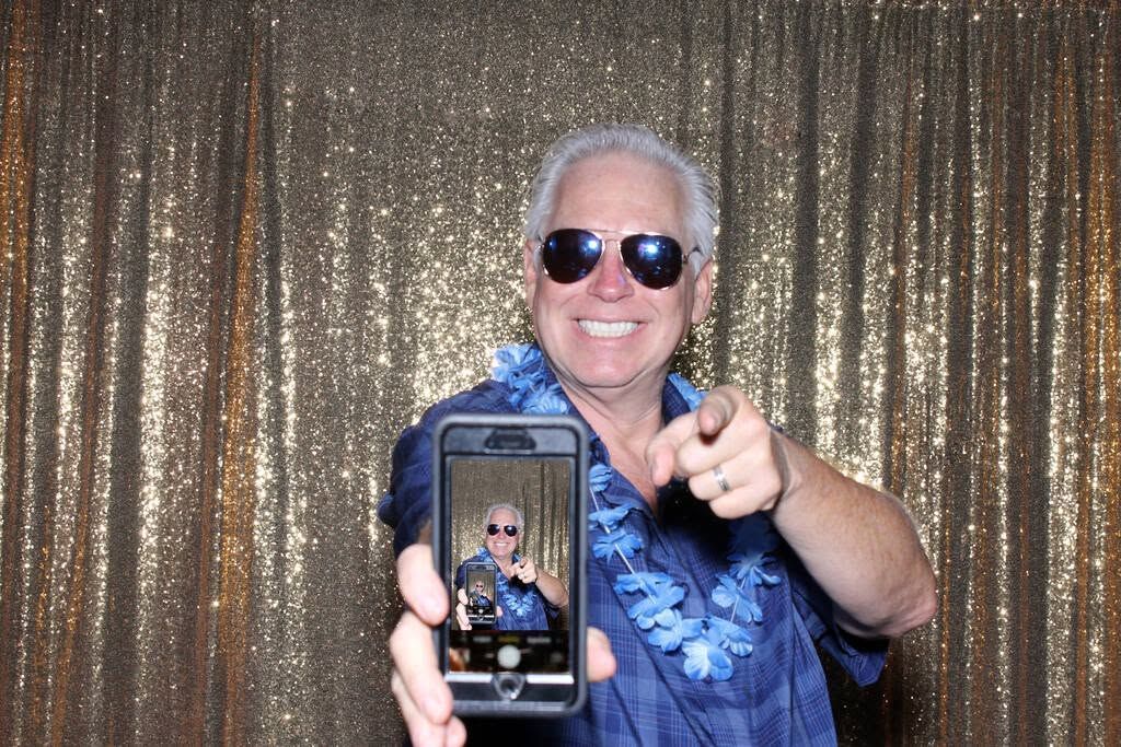 Smiling person in sunglasses holding a phone for a selfie in front of a gold sequin backdrop