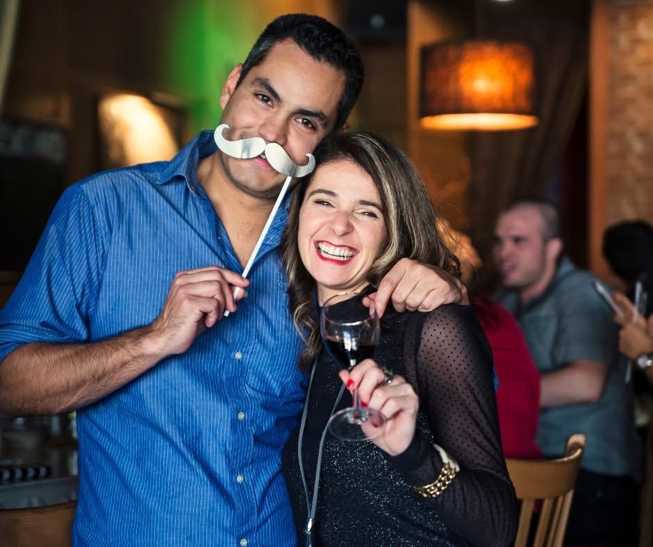 Two people posing at a bar, smiling and holding drinks, with a fake mustache prop between them.