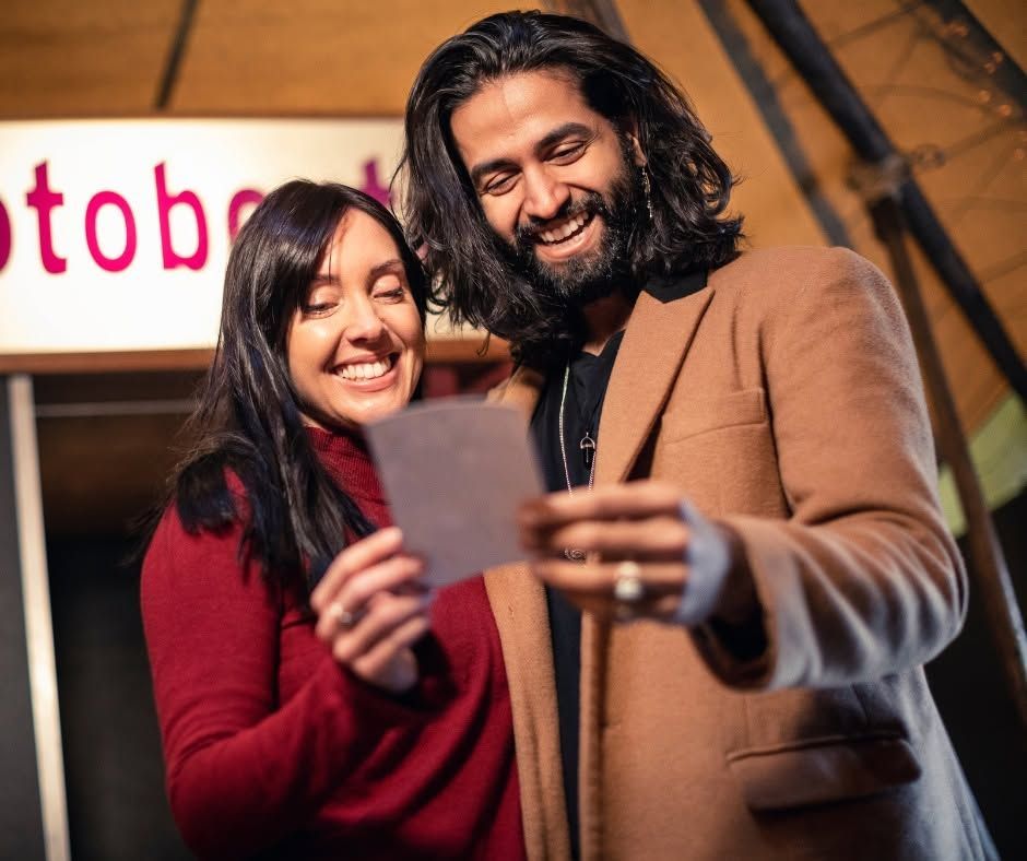 Two smiling people in coats looking at a photo together indoors
