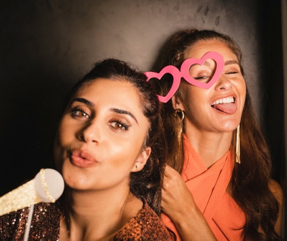 Two women posing at a party, one blowing a kiss and the other laughing, with pink heart glasses overlay