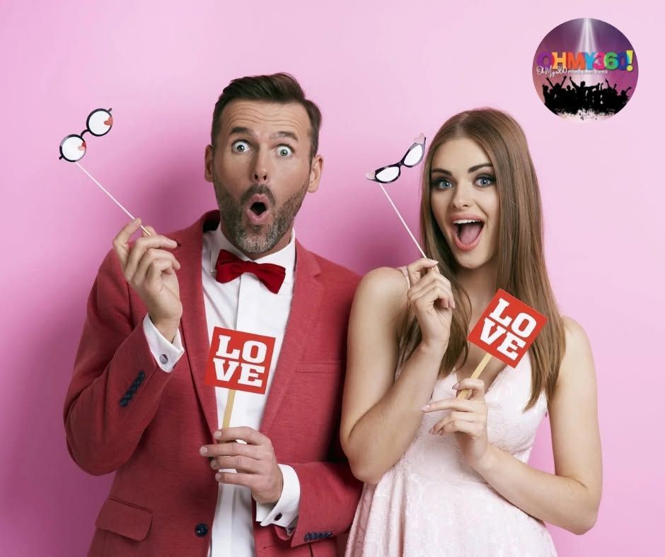 Two smiling people in pink studio outfits holding heart props and “LOVE” cards against a pink background