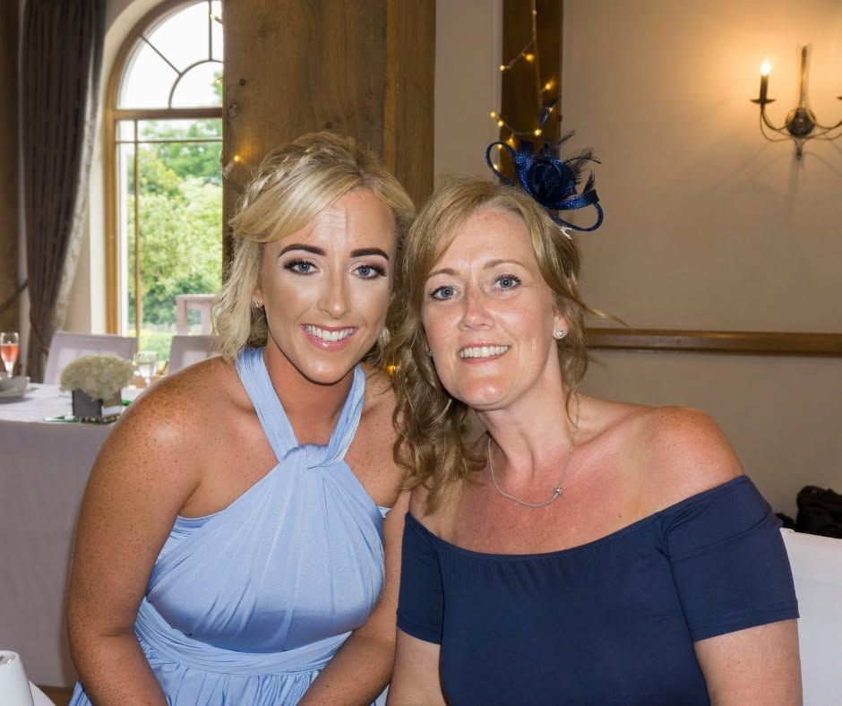 Two people smiling at a formal indoor event, wearing blue dresses, seated by a window.