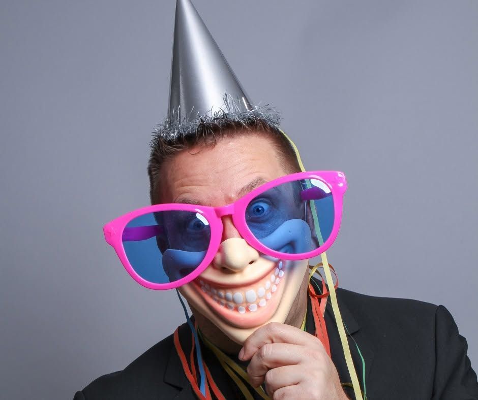 Person in a silver party hat and pink sunglasses grinning against a gray background.