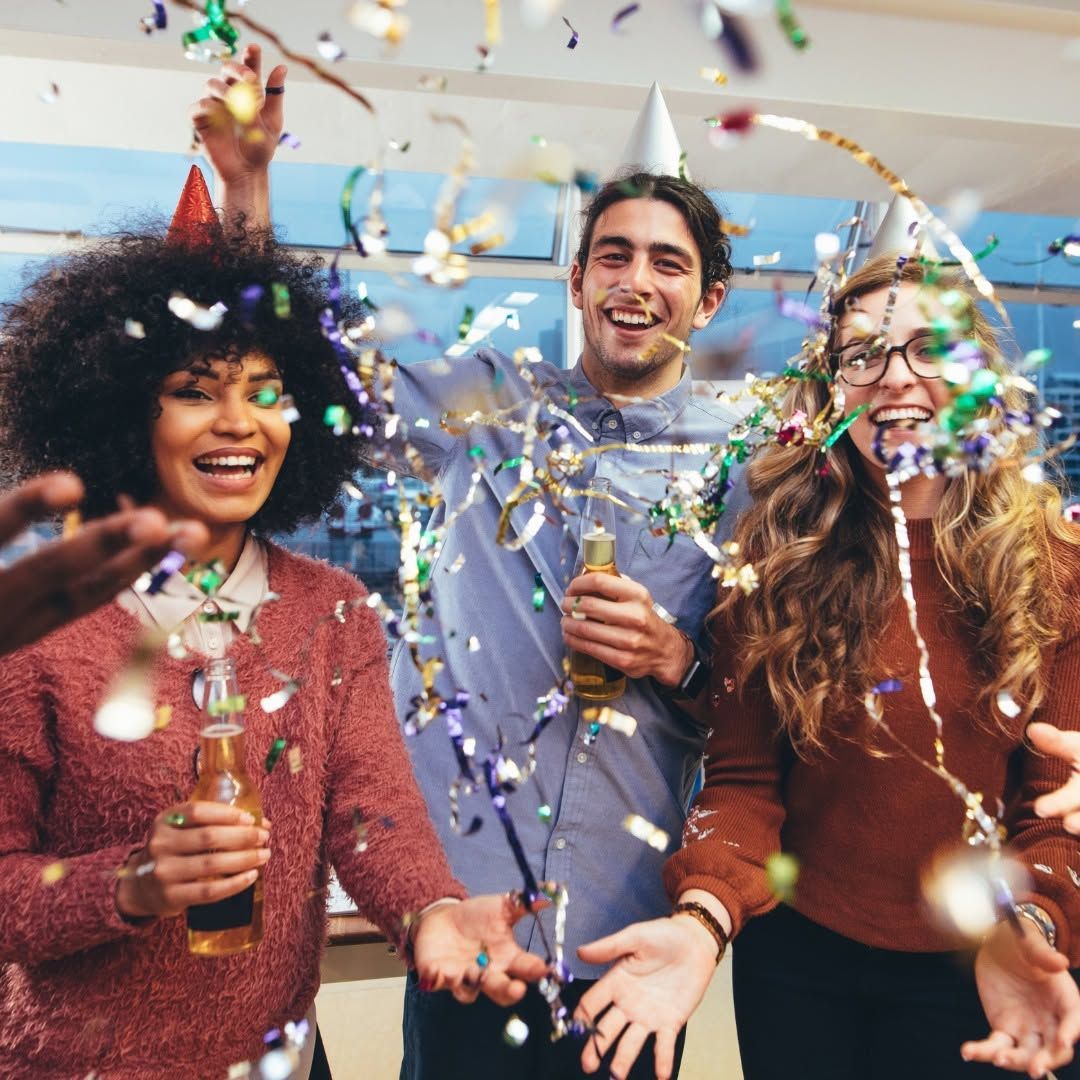 Three friends celebrating with confetti and party poppers, smiling in a bright indoor setting.