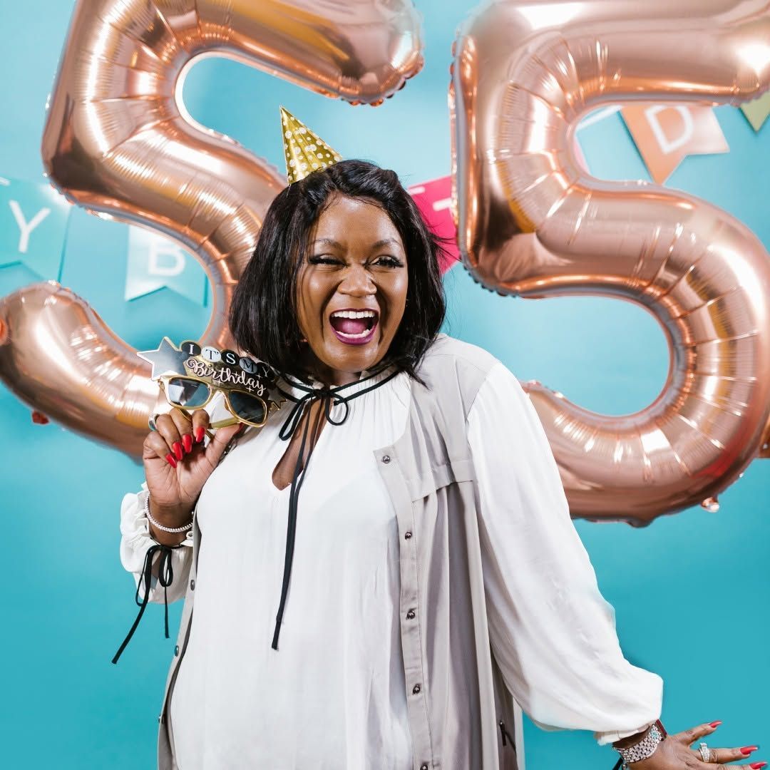 Woman in white dress celebrating with a party horn, gold party hat, and rose gold balloons behind her