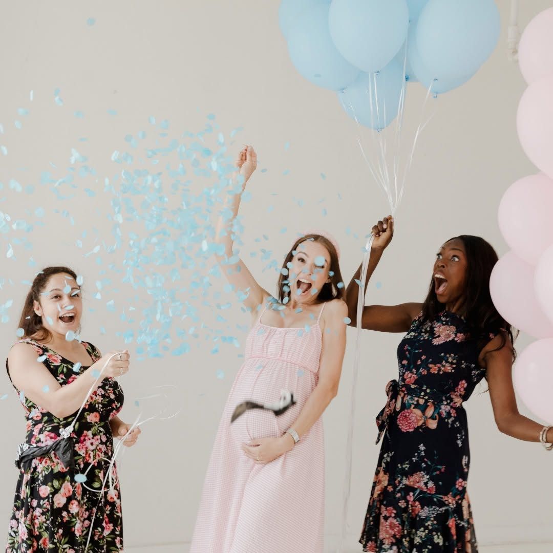 Three friends in floral and pastel dresses celebrate with blue balloons and confetti indoors