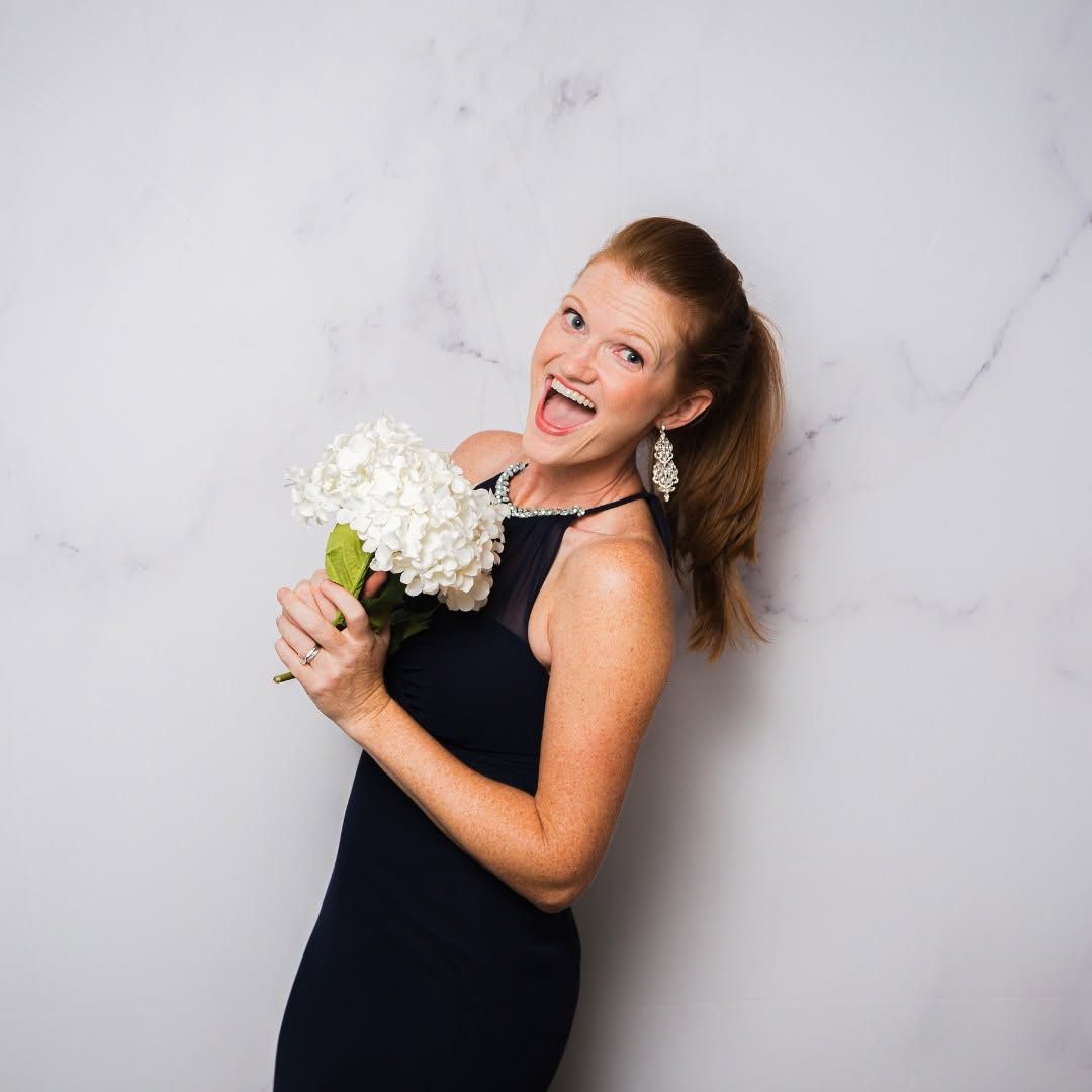 Smiling woman in a black dress holding a white bouquet against a light gray wall