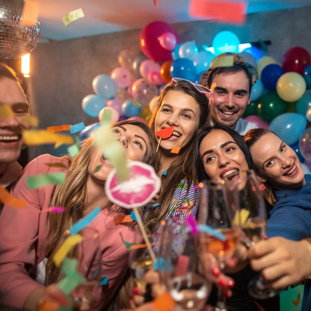 Friends celebrating at a party with balloons, confetti, and drinks, smiling in a festive indoor setting