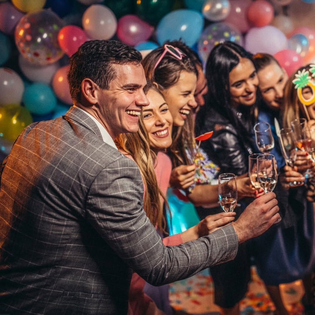 Friends smiling and toasting drinks at a colorful party with balloons and confetti