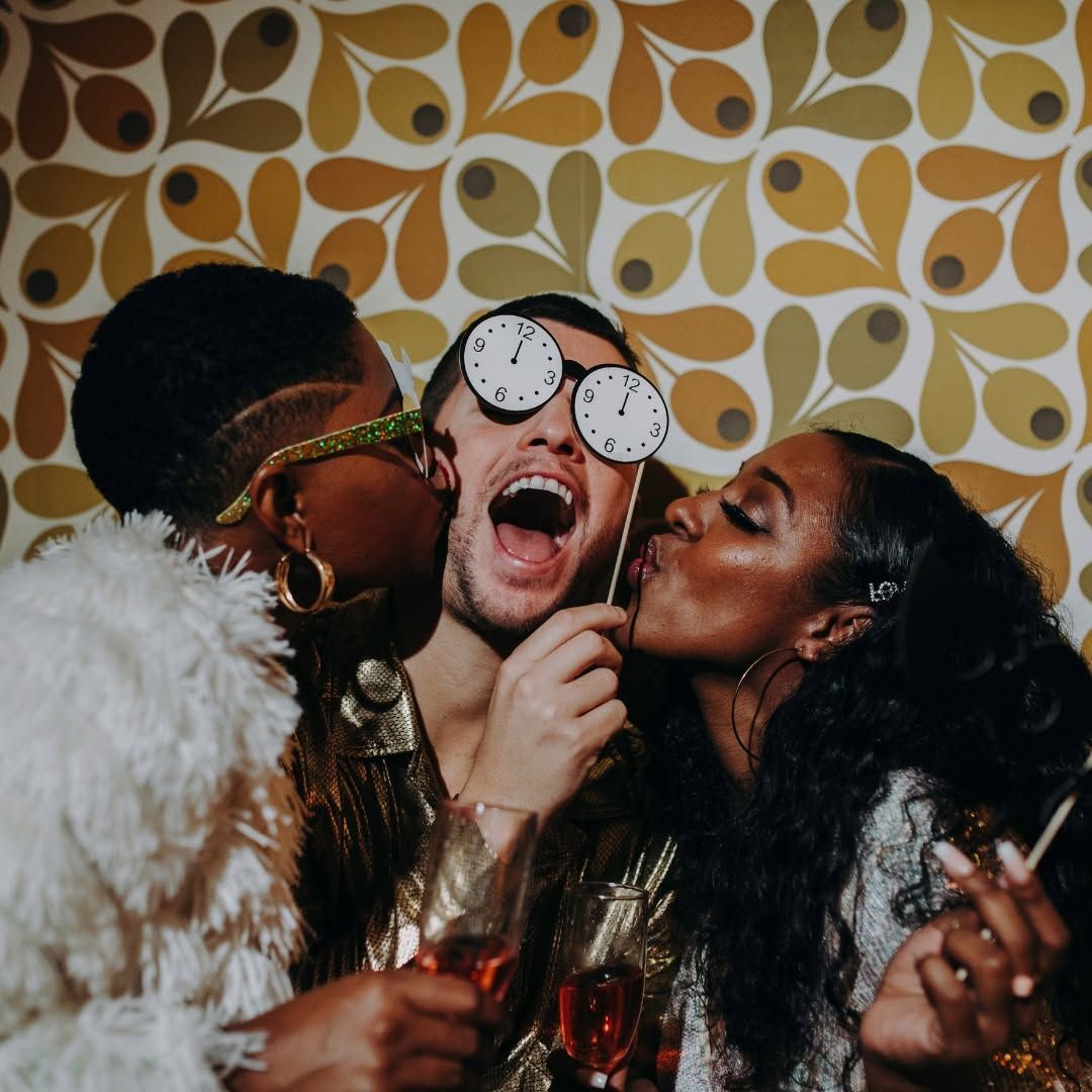 Friends in festive attire celebrate with champagne, kissing a man wearing clock-shaped sunglasses.
