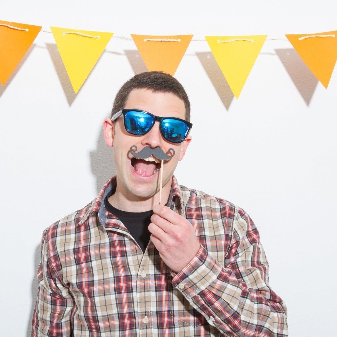Person in blue sunglasses and plaid shirt posing with a fake mustache under yellow party bunting