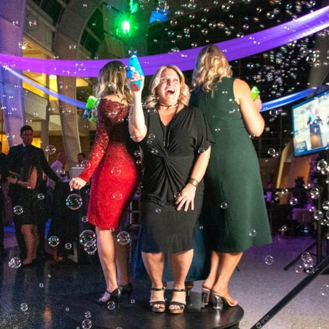 Three women in dresses dancing at a party under colorful lights, bubbles floating around them.