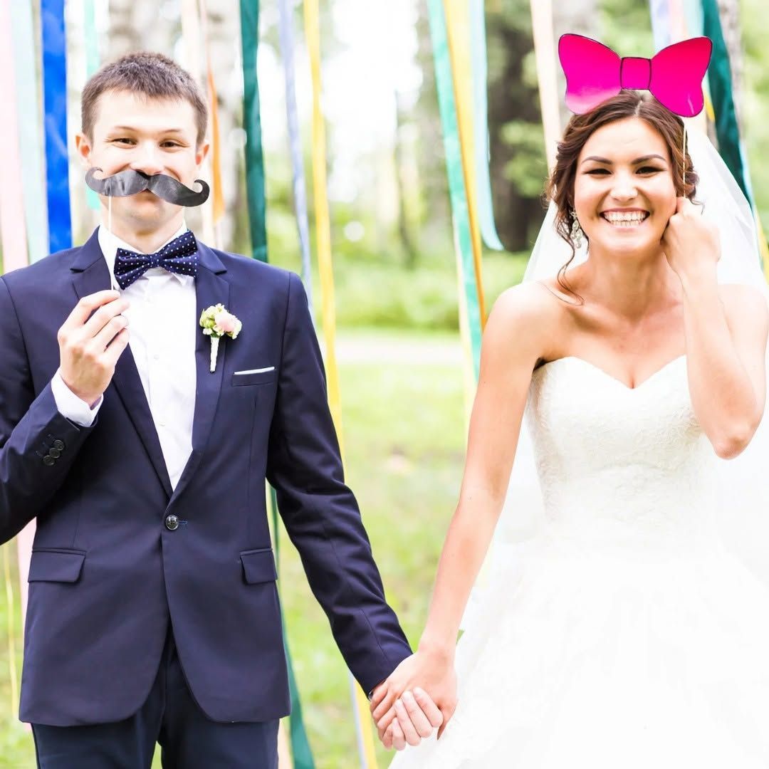 Bride and groom in wedding attire holding hands outdoors, both smiling with playful fake mustaches