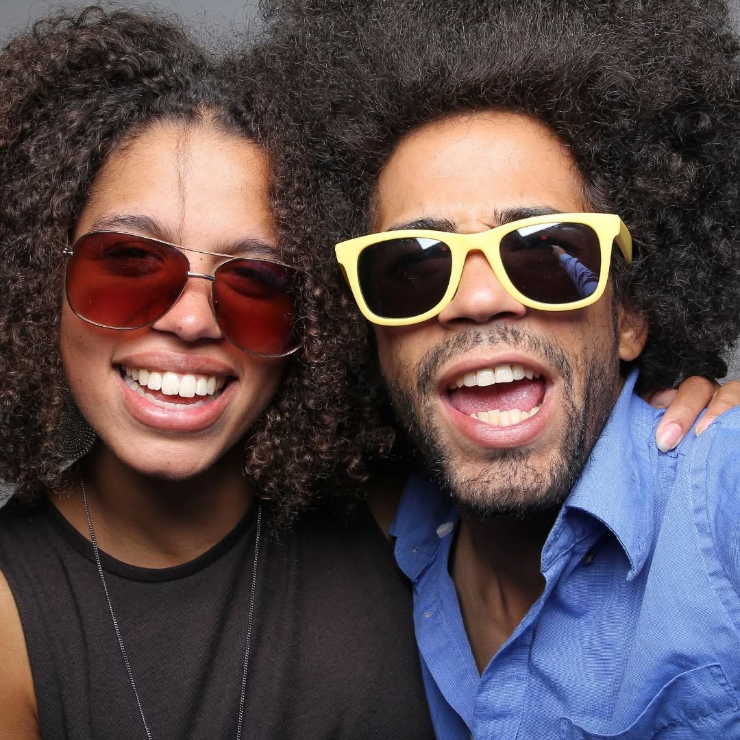 Two smiling people wearing sunglasses posing close together against a dark background
