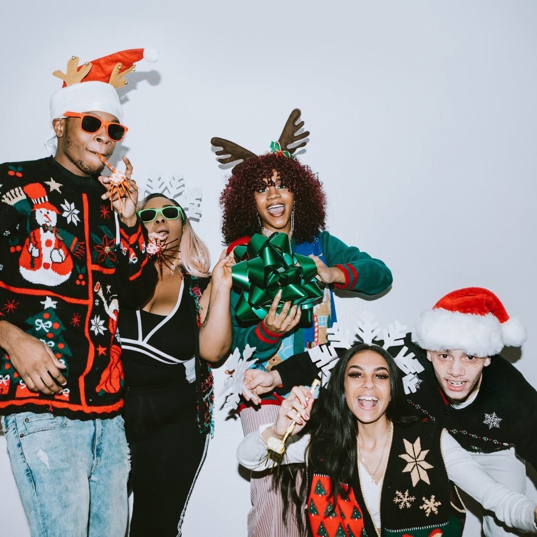 Five people in festive holiday sweaters and hats pose together, smiling against a plain light background.