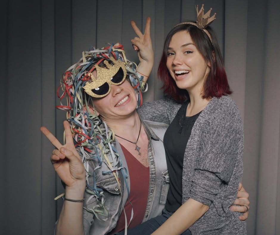 Two smiling people posing with playful props, one wearing a colorful wig and sunglasses, against gray curtains