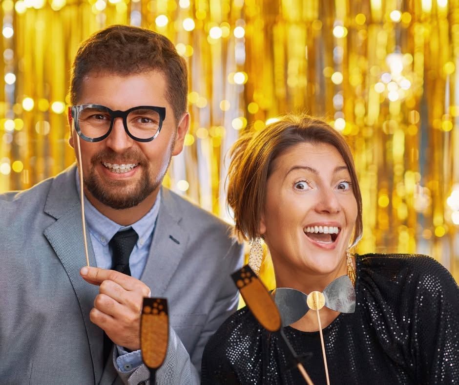 Two smiling people in formalwear holding novelty microphones in front of gold tinsel curtains