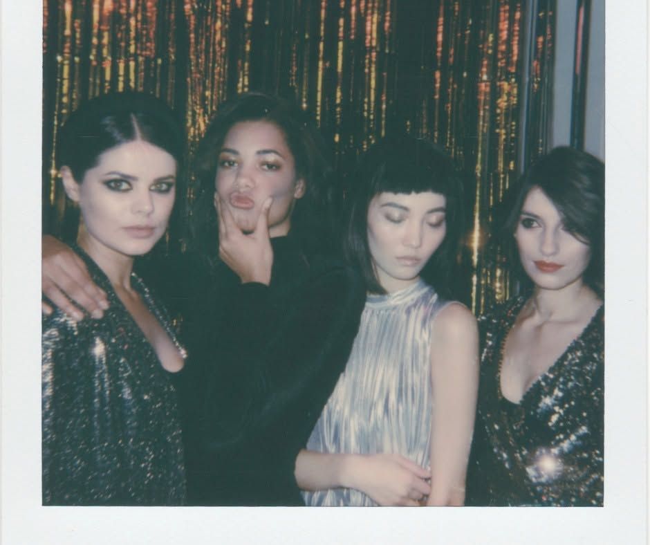 Four women posing before a shimmering black-and-gold curtain, wearing dark evening outfits.