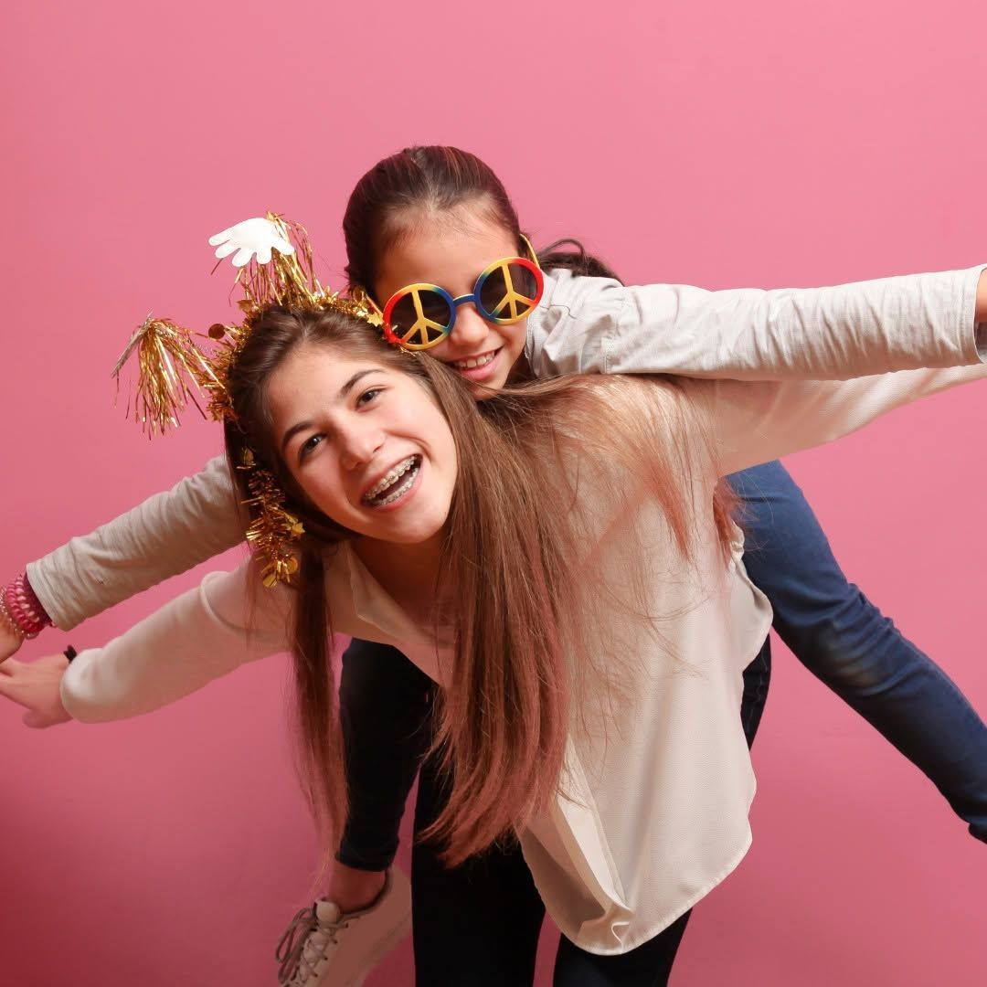 Two smiling friends posing playfully against a pink backdrop, one wearing gold party headpieces and sunglasses