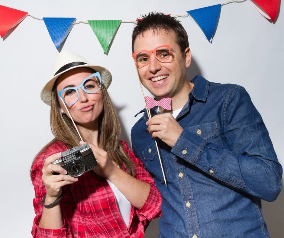 Two people posing with cameras and props under colorful bunting at a party backdrop