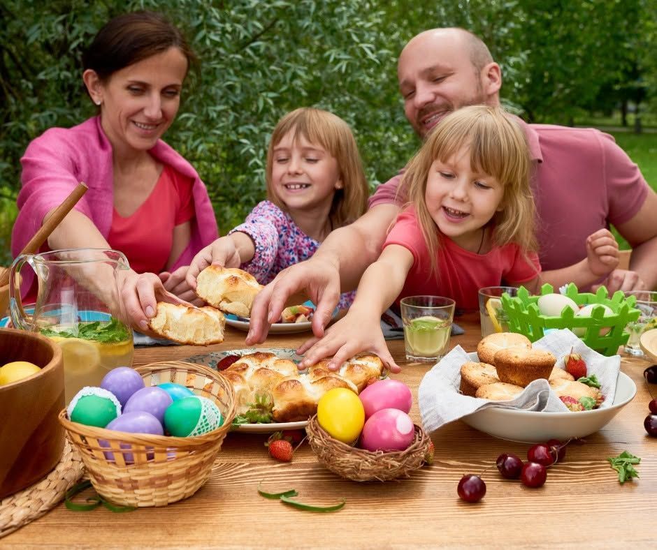 Family decorating Easter eggs outdoors at a table with bread, fruit, and flowers.