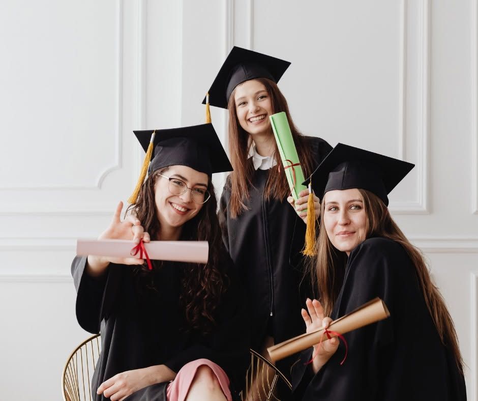 Three graduates in black caps and gowns pose with diplomas, smiling indoors.