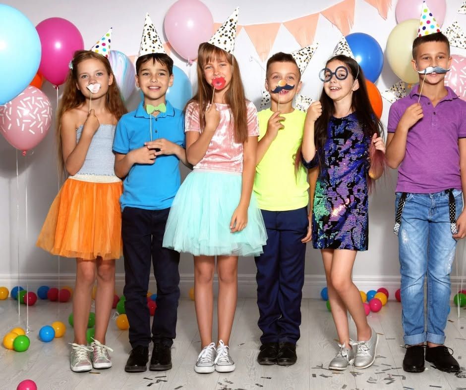 Six children in party hats stand together smiling at a birthday party with balloons and confetti decorations