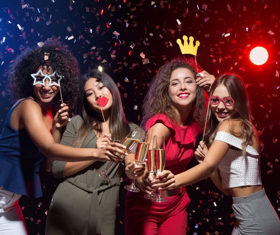 Four friends in party attire toast with drinks amid confetti and colorful lights