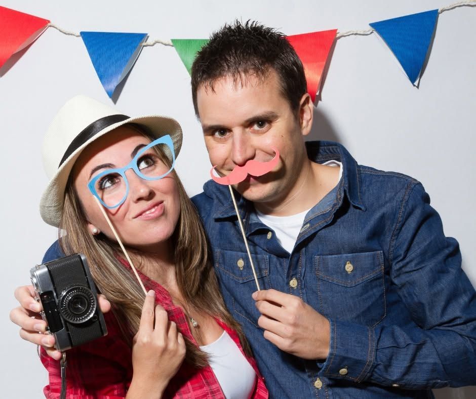 Two people posing with props at a party, holding a camera and wearing playful hats and mustaches.