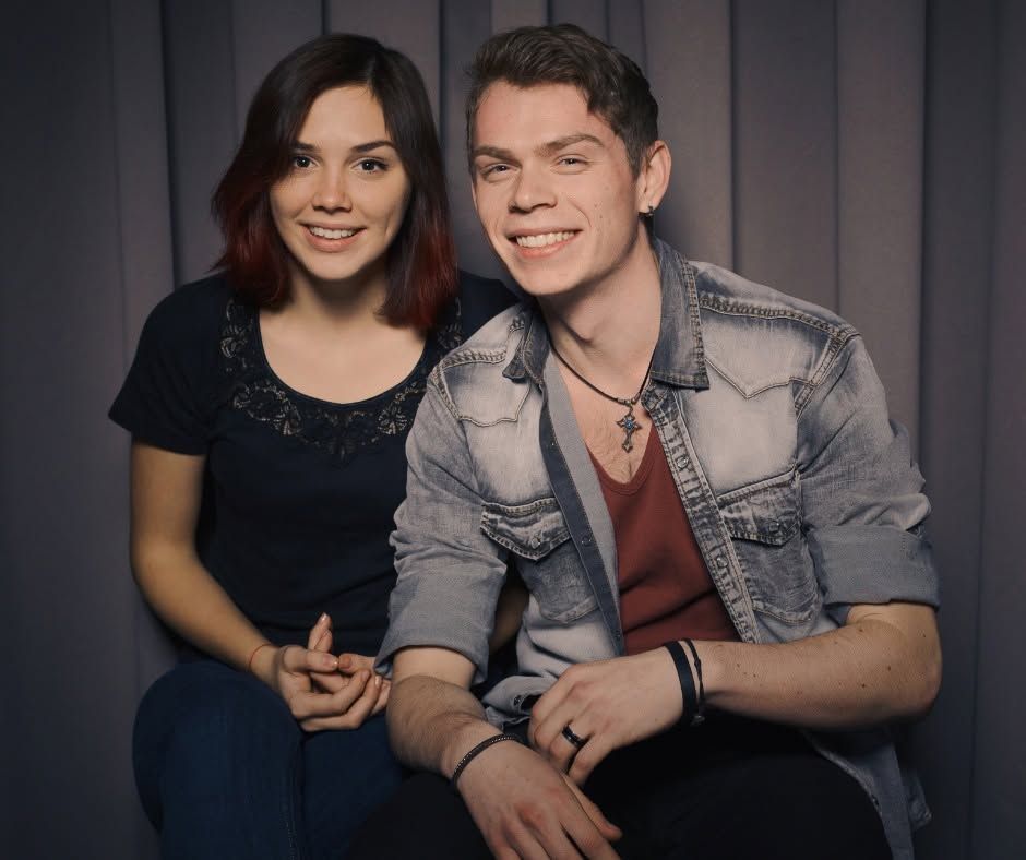 Two smiling people seated against a gray backdrop, one in a dark top and one in a light denim jacket.