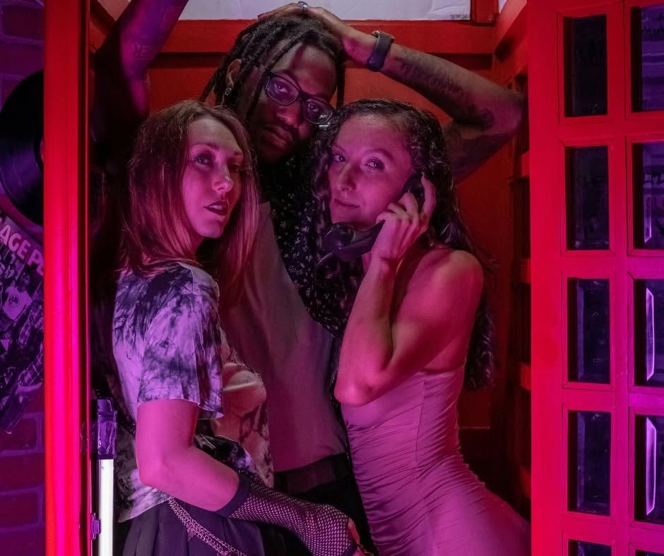 Three people pose closely in a neon pink-lit booth with red-framed windows.
