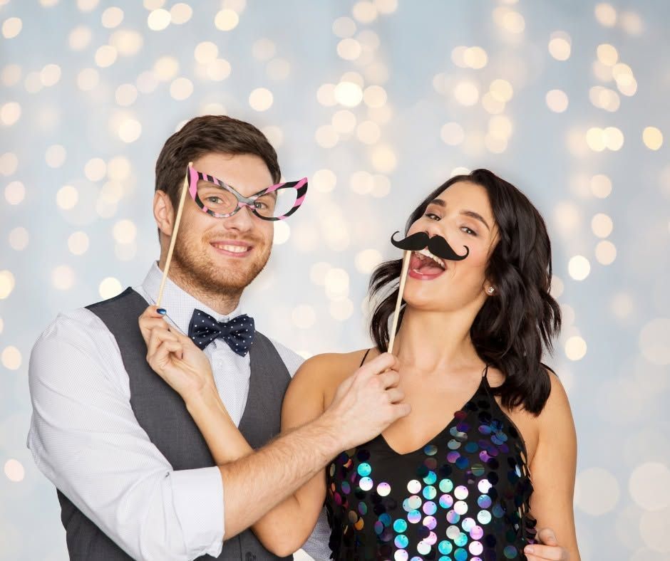 Two people in party props pose in front of a sparkling light backdrop, smiling and playful.