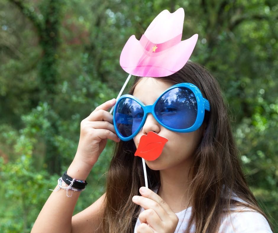 Person outdoors wearing oversized blue glasses and a pink flower hat prop, holding a red party prop.